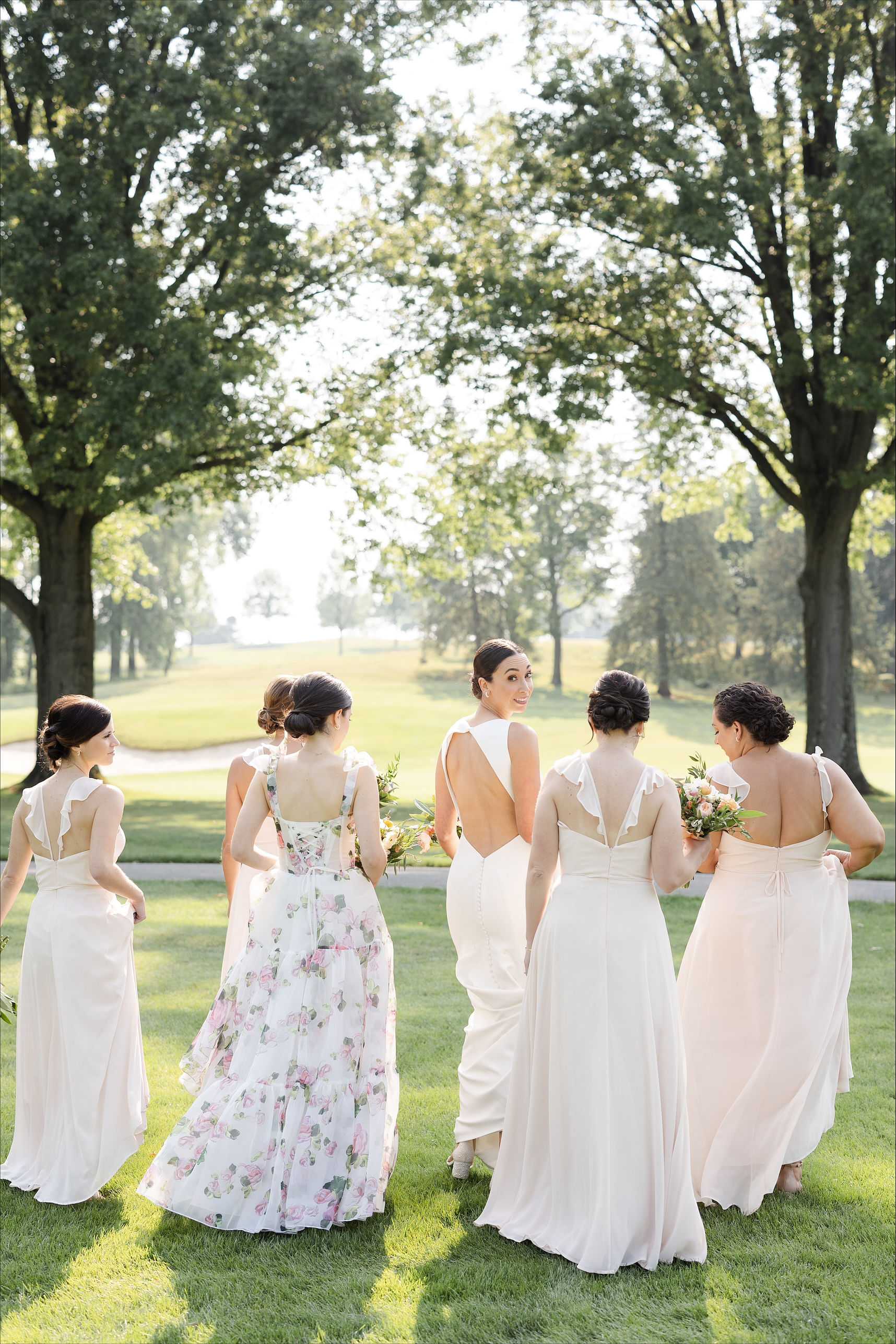 Bridesmaids in pastel dresses holding bouquets, walking together on a lush green lawn.